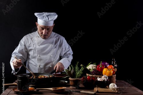 Male cook in white uniform and hat with ladle mixes the ingredients onto the cooking pan photo