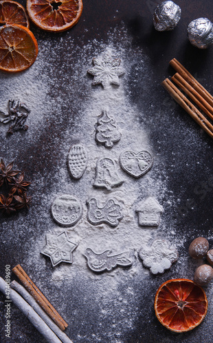 Christmas gingerbread cookies on a dark background, white flour in fir tree shape, dry oranges, spices and rolling pin, flat lay