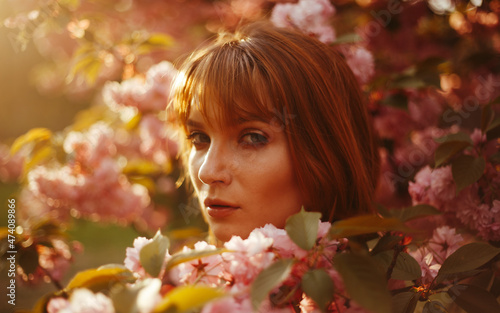 a red-haired girl with fringe looks curiously while standing between the flowers of a blossoming tree