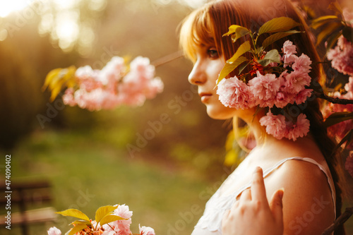 a red-haired girl with fringe looks curiously while standing between the flowers of a blossoming tree