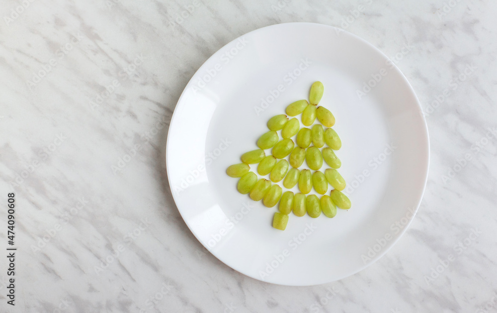Funny edible Christmas tree, breakfast or snack idea for kids. Made of grape. decor with garaland and dry orange. New Year concept. White marble background. top view, flat lay, space for text Funny edible Christmas tree, breakfast or snack idea for kids. Made of grape. decor with garaland and dry orange. New Year concept. White marble background. top view, flat lay, space for text