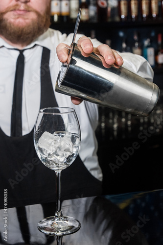 Professional bartender with a beard pours liquid into a glass of ice cubes from a tool for mixing and preparing alcoholic beverages cocktails metal shaker