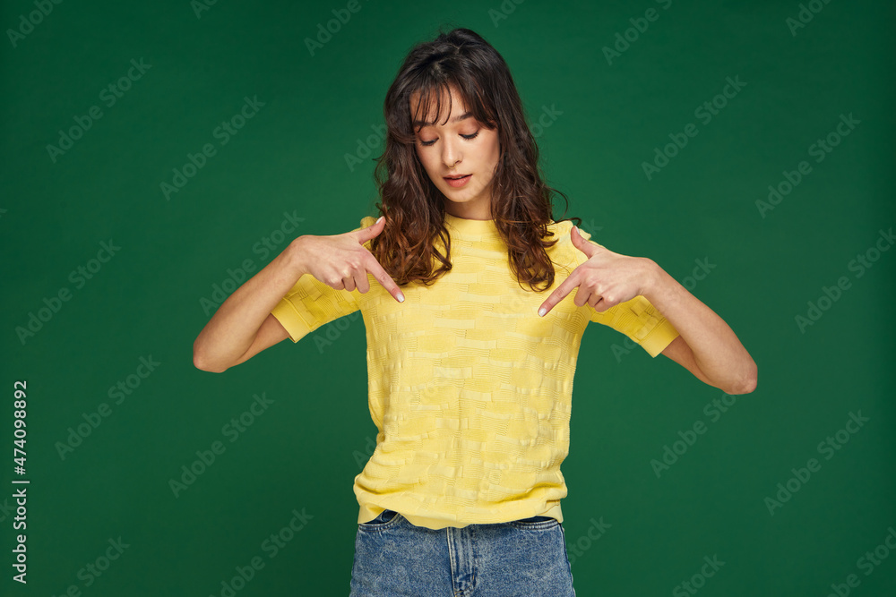 Modern beautiful young girl pointing down by fingers on link to subscribe on green studio background. Good sale offer 