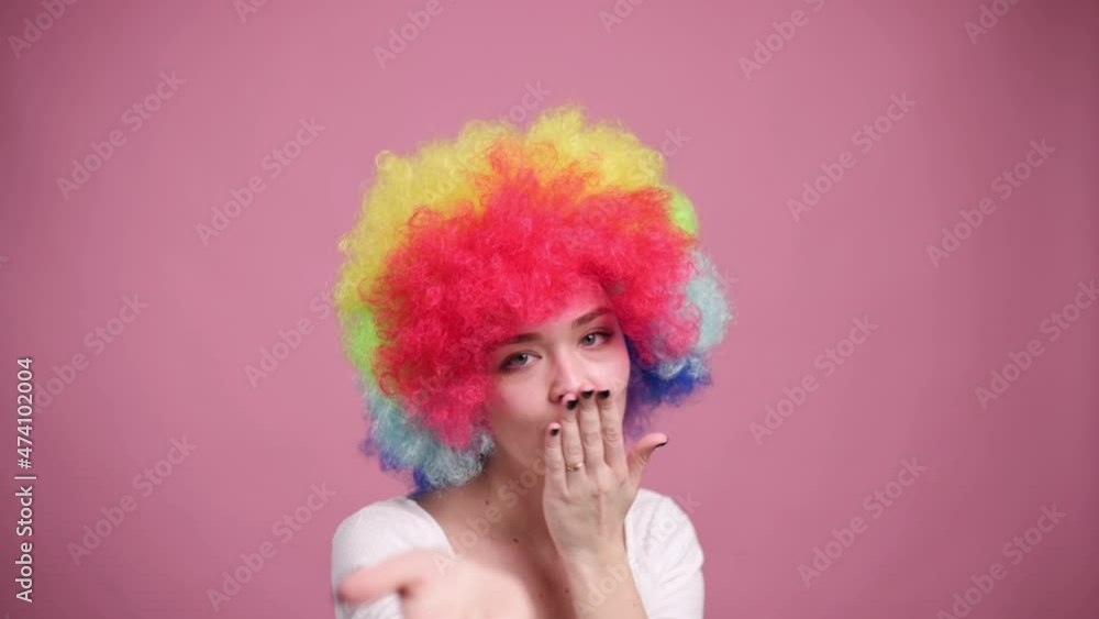 Smiling young attractive female in colorful wig sends air kisses on pink background. Close-up. Smiling young attractive female in colorful wig sends air kisses on pink background. Close-up.