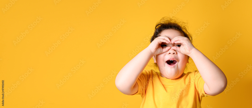 Little Asian girl makes glasses from fingers and smiles, looks at the camera. Yellow background. Blank Space Little Asian girl makes glasses from fingers and smiles, looks at the camera. Yellow background. Blank Space