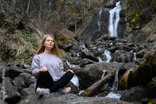 Girl sits on a large stone and meditates near a waterfall next to a mountain river.