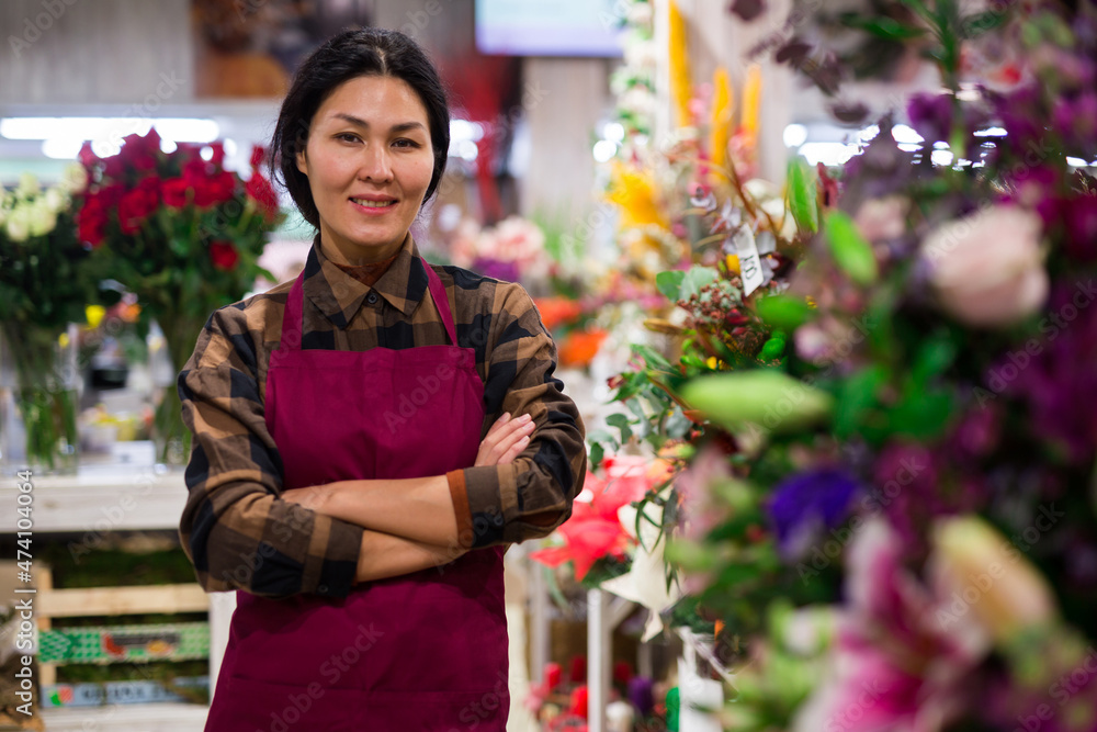 Asian woman florist in uniform standing among shelves with plants and flowers, smiling and looking in camera..