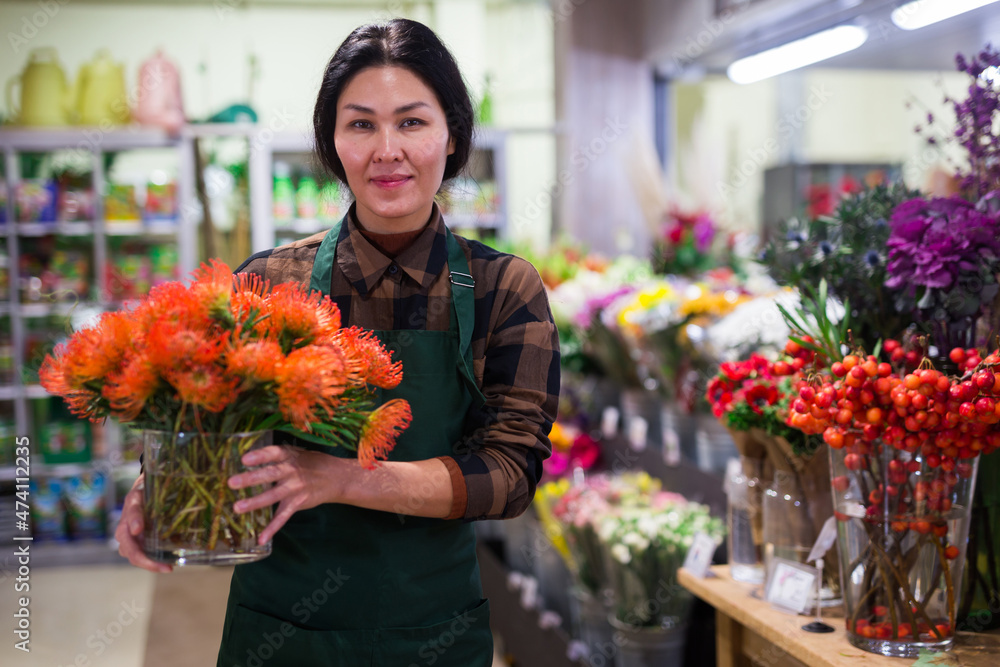 Asian woman florist carrying glass vase with orange flowers in salesroom of floral shop.