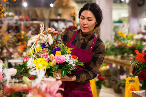 Asian woman florist carrying basket with flowers in salesroom of floral shop.