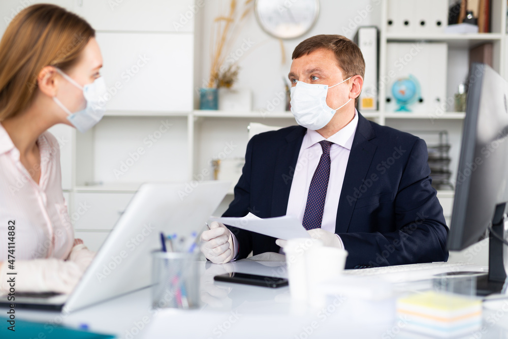 Fototapeta premium Portrait of busy entrepreneur in medical face mask and latex gloves working with female coworker in office. Concept of precautions and social distancing in coronavirus pandemic..
