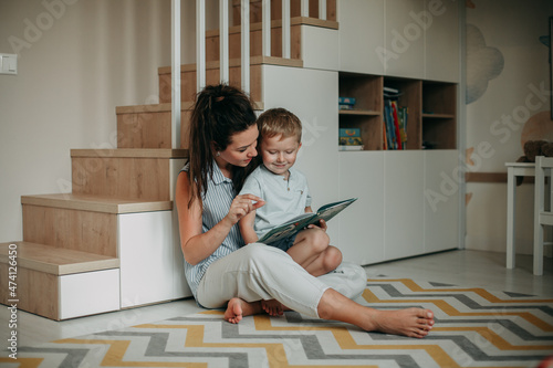 A young girl of 30 years old brunette with long hair in a white shirt with a naked strip and in white jeans at home in the nursery room sits on the floor with her son and reads a book to him.