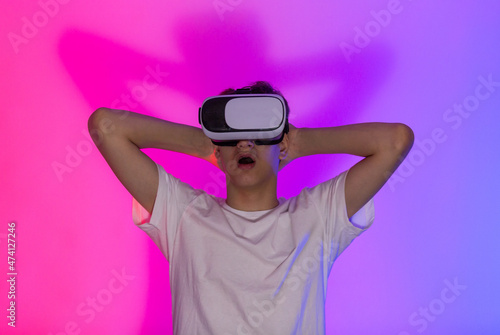 Cute teenager in white t shirt smiling while wearing modern VR headset, standing on pink purple background. Modern hobby, playing video games for children, teens