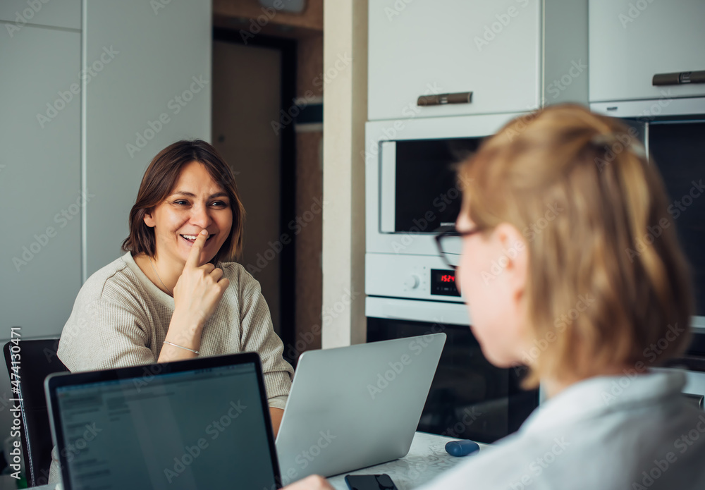 Coworking, remote work, home office, startup. Two young pretty women sitting with laptops facing each other, focus on the brunette's face. Coworking, remote work, home office, startup. Two young pretty women sitting with laptops facing each other, focus on the brunette's face.