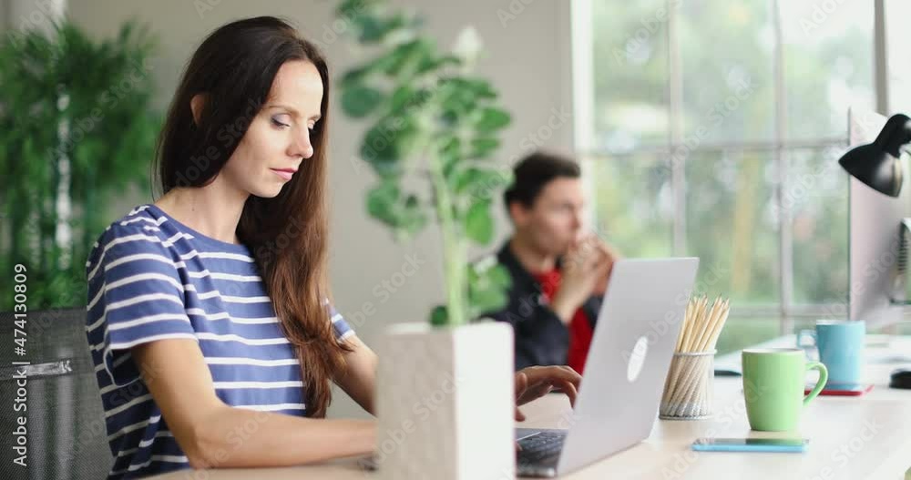 Millennial Caucasian young professional success female worker in casual outfit sitting working typing laptop notebook computer on table with tree pot coffee cup pencils and lamp with male colleague. Millennial Caucasian young professional success female worker in casual outfit sitting working typing laptop notebook computer on table with tree pot coffee cup pencils and lamp with male colleague.