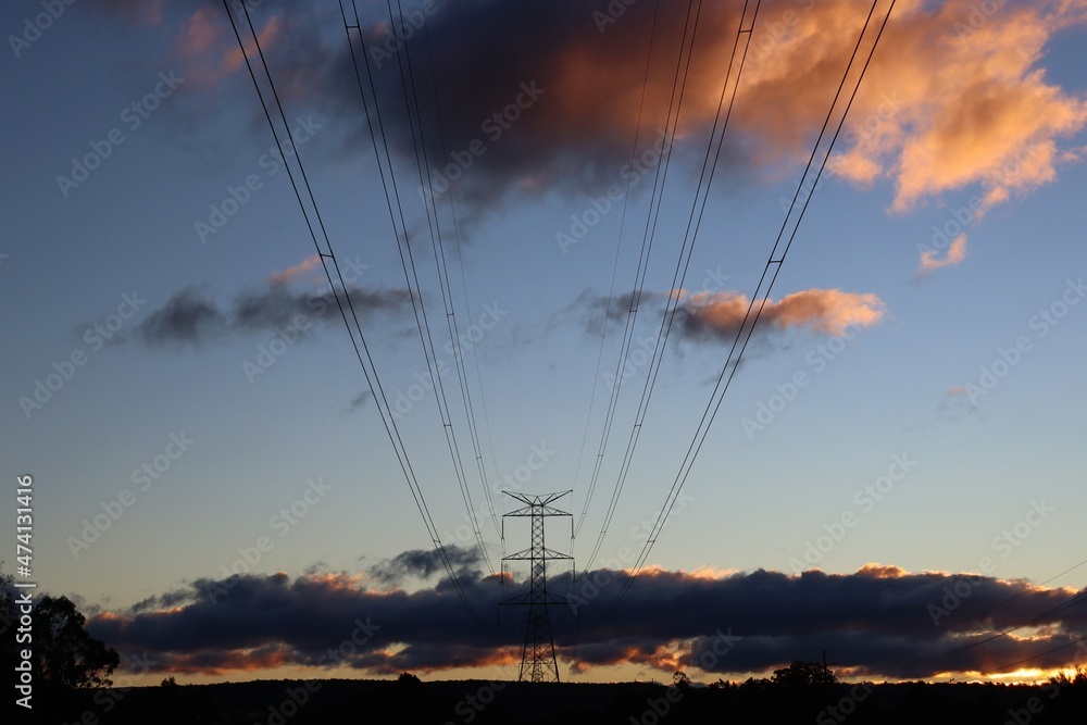 Large overland power lines spanning across suburban wetlands area toward mountains in the distance, with the sun setting in the background.