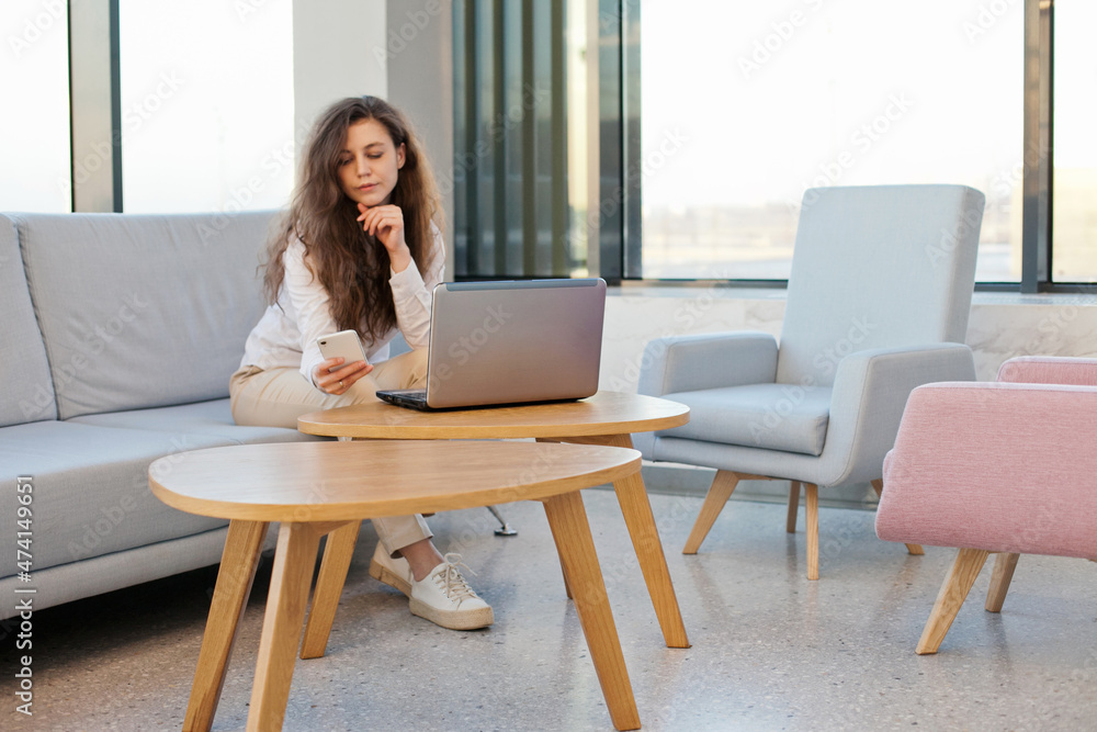 woman sitting on a sofa or couch working on a laptop with mobile phone in the office with big windows woman sitting on a sofa or couch working on a laptop with mobile phone in the office with big windows