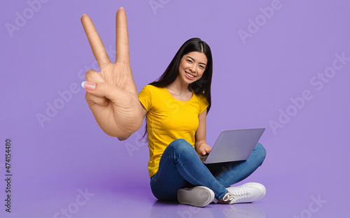 Remote job or education. Smiling young Asian woman sitting with laptop, showing big peace or victory gesture © Prostock-studio