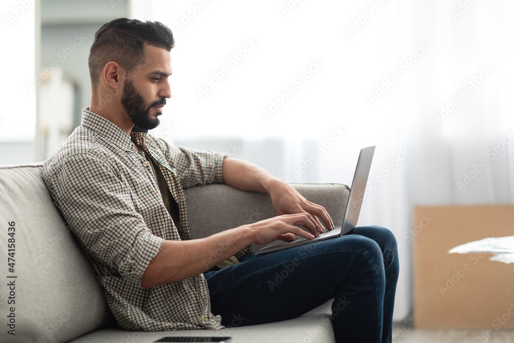 Online work, communication. Young Arab guy making video call, browsing internet on laptop, sitting on couch at home