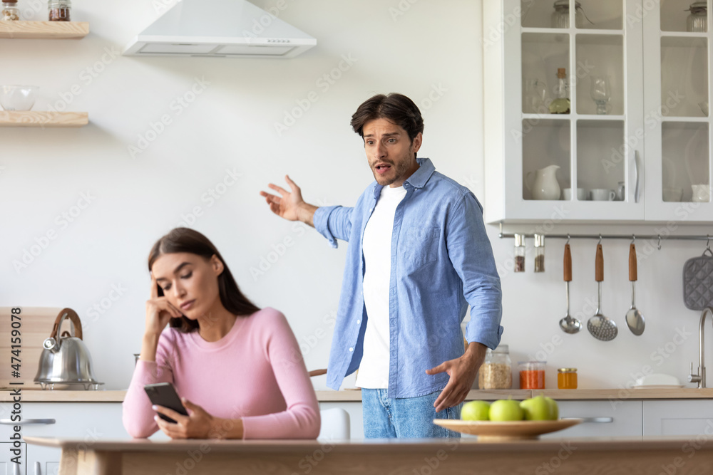 Sad millennial man scolds wife, yelling at woman with smartphone on kitchen interior during covid-19 Sad millennial man scolds wife, yelling at woman with smartphone on kitchen interior during covid-19