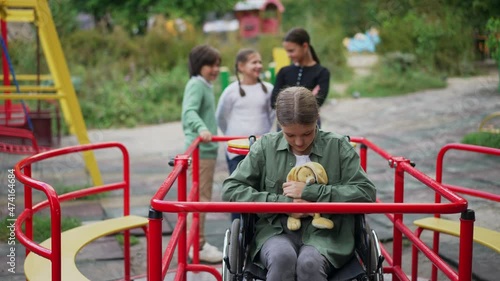 Portrait of sad Caucasian disabled girl in wheelchair hugging toy and looking at camera in slow motion as group of children standing at background talking laughing. Depressed child on playground photo