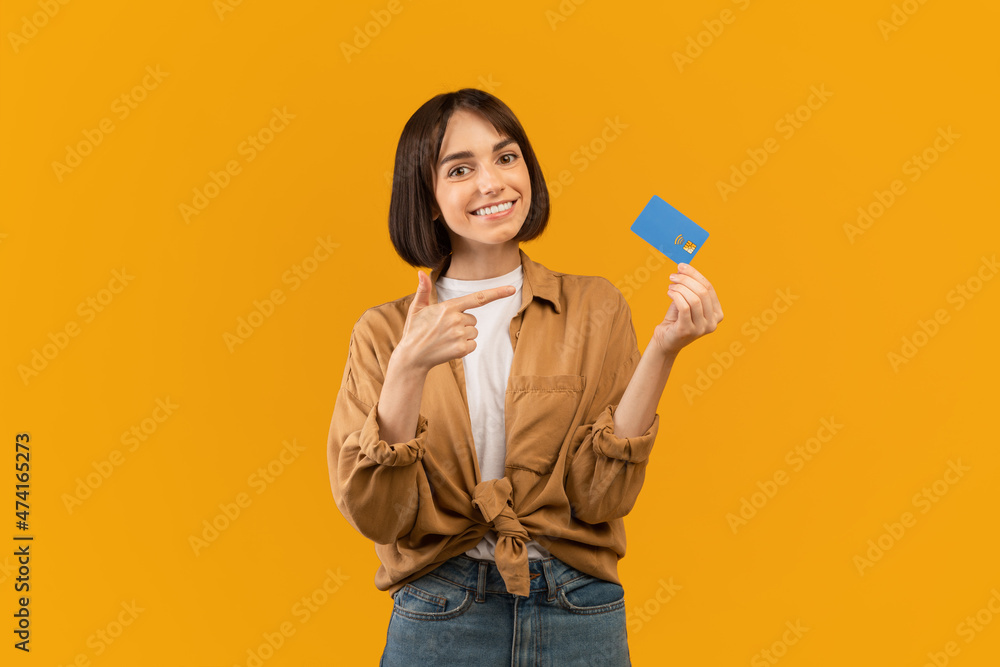 Positive young lady pointing at credit card recommending banking service, posing over yellow background and smiling Positive young lady pointing at credit card recommending banking service, posing over yellow background and smiling