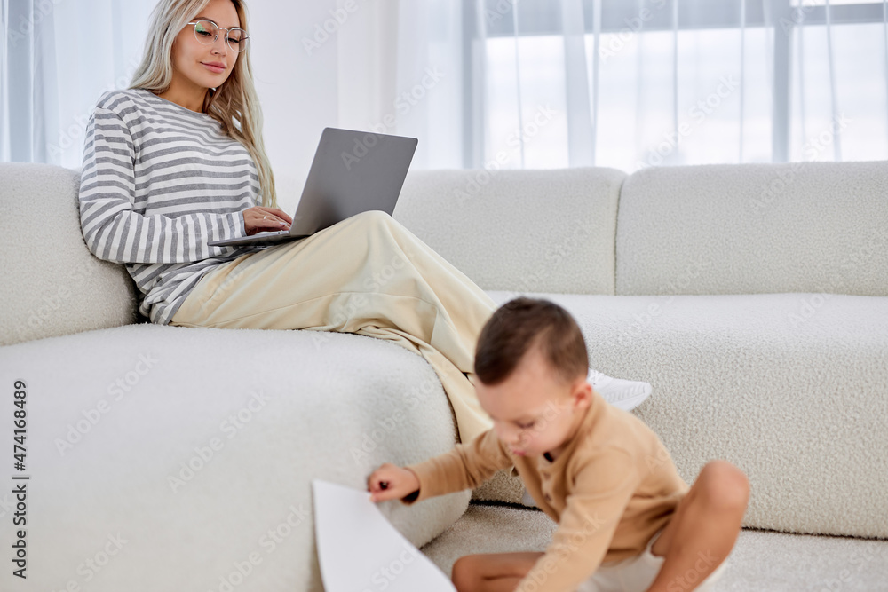 young blonde caucasian woman sitting on sofa working on laptop while child boy son is playing on floor. Office at home, Freelance and parenting concept. in bright cozy room. at home young blonde caucasian woman sitting on sofa working on laptop while child boy son is playing on floor. Office at home, Freelance and parenting concept. in bright cozy room. at home