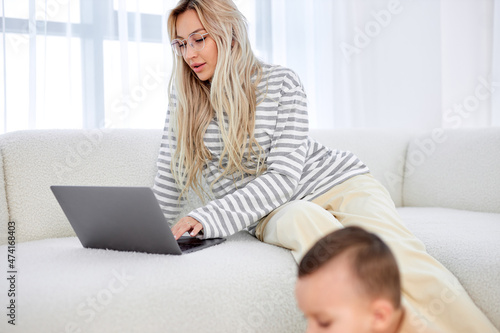 blonde caucasian mom working at home on sofa writing an email while child playing on floor. Young pretty woman working from home, while in quarantine isolation during the Covid-19 health crisis
