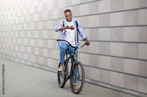 Happy African American guy with earphones listening to music and checking time on smartwatch during bike ride outside