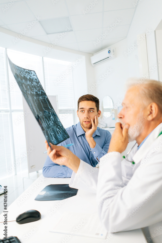 Obraz premium Vertical shot of mature adult male physician consulting scared young man patient giving bad news explaining results of MRI image. Unhappy frustrated young man listening to bad news, looking at camera.
