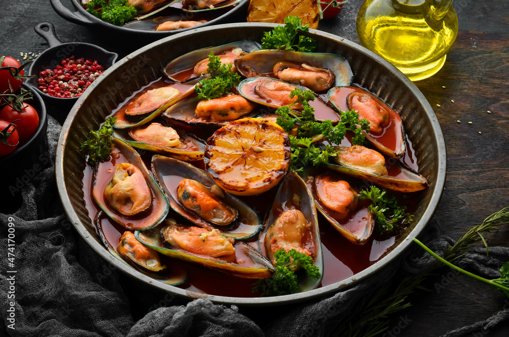 Metal tray with mussels cooked in tomato sauce with garlic, parsley and lemon. On a black stone background.