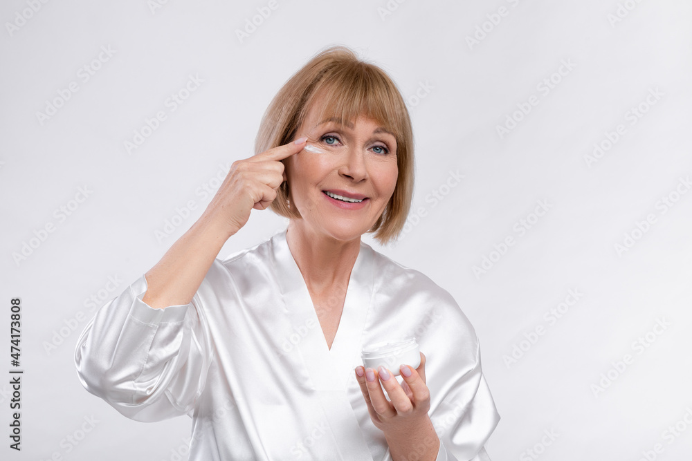 Sensual senior lady in silk bath robe applying face cream and smiling at camera over light studio background Sensual senior lady in silk bath robe applying face cream and smiling at camera over light studio background