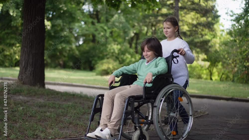Wide shot smiling disabled boy with girl pushing wheelchair in slow motion outdoors. Happy Caucasian brother and sister enjoying leisure in summer park. Family support and disability concept Wide shot smiling disabled boy with girl pushing wheelchair in slow motion outdoors. Happy Caucasian brother and sister enjoying leisure in summer park. Family support and disability concept