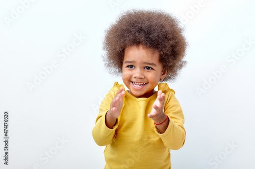Excited black fluffy child girl clapping and applauding, happy and joyful, smiling. Adorable charming kid in yellow shirt have fun isolated on white studio background. Portrait, copy space.