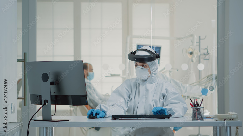 Portrait of dental assistent typing on computer keyboard wearing protection uniform with face shield, mask, gloves and coverall. Stomatology nurse using monitor technology at oral clinic Portrait of dental assistent typing on computer keyboard wearing protection uniform with face shield, mask, gloves and coverall. Stomatology nurse using monitor technology at oral clinic