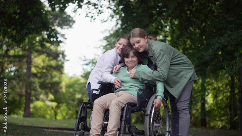Wide shot portrait happy smiling boy in wheelchair with girls hugging friend. Positive Caucasian disabled brother with sisters in summer park posing in slow motion. Childhood and support concept Wide shot portrait happy smiling boy in wheelchair with girls hugging friend. Positive Caucasian disabled brother with sisters in summer park posing in slow motion. Childhood and support concept