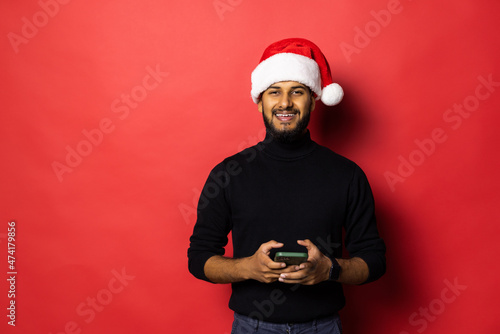 Young indian man in Santa hat use phone send greeting online standing isolated on red background photo