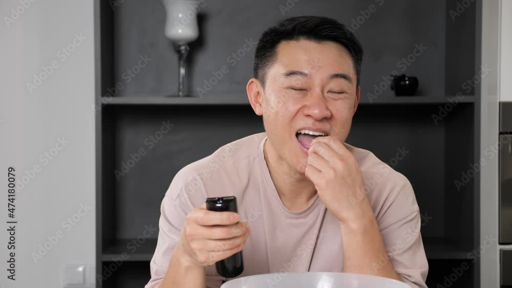 Open-eyed young asian man watching TV and eating pop corn standing in the kitchen at home. Close up of male shows wow emotion while watching movie or tv show. Open-eyed young asian man watching TV and eating pop corn standing in the kitchen at home. Close up of male shows wow emotion while watching movie or tv show.