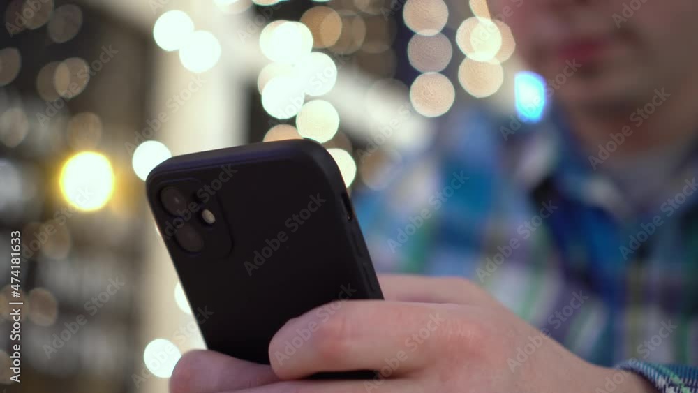 A young man is sitting at a table in a cafe in the evening and texting on the phone. A man with a phone in his hands on the background of garlands. Close-up. A young man is sitting at a table in a cafe in the evening and texting on the phone. A man with a phone in his hands on the background of garlands. Close-up.