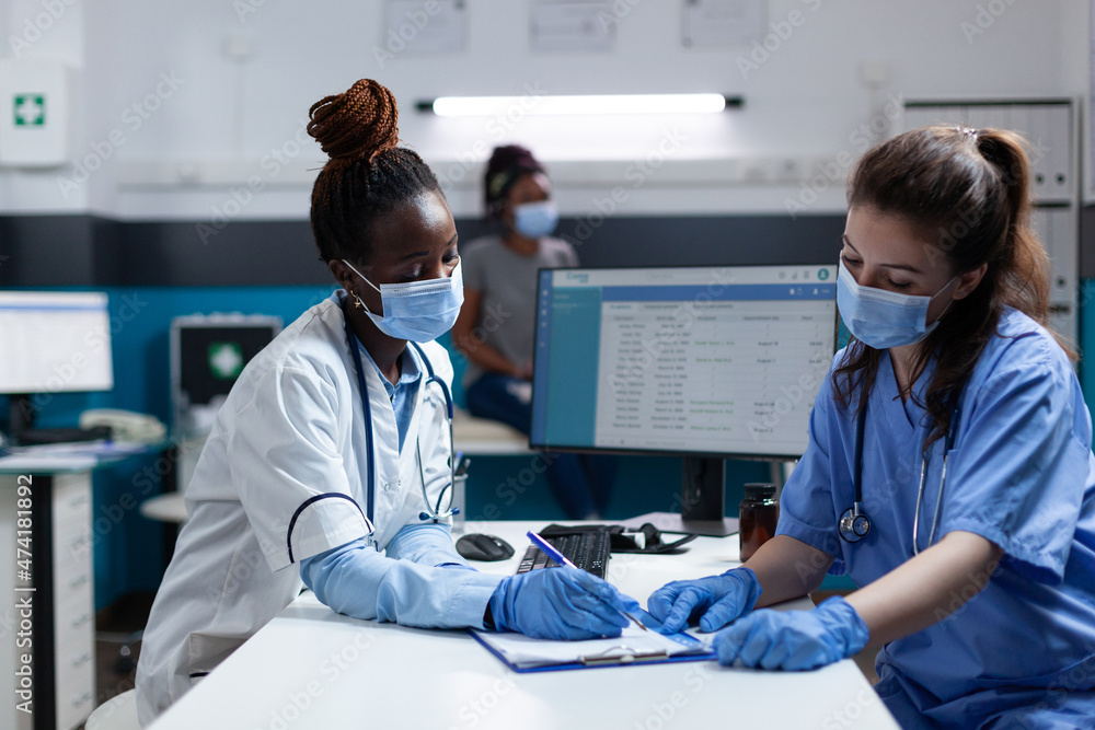 Obraz premium Medical team with protective face mask to prevent infection with covid19 writing medication prescription during clinical appointment in hospital office. African american doctor examining sick patient