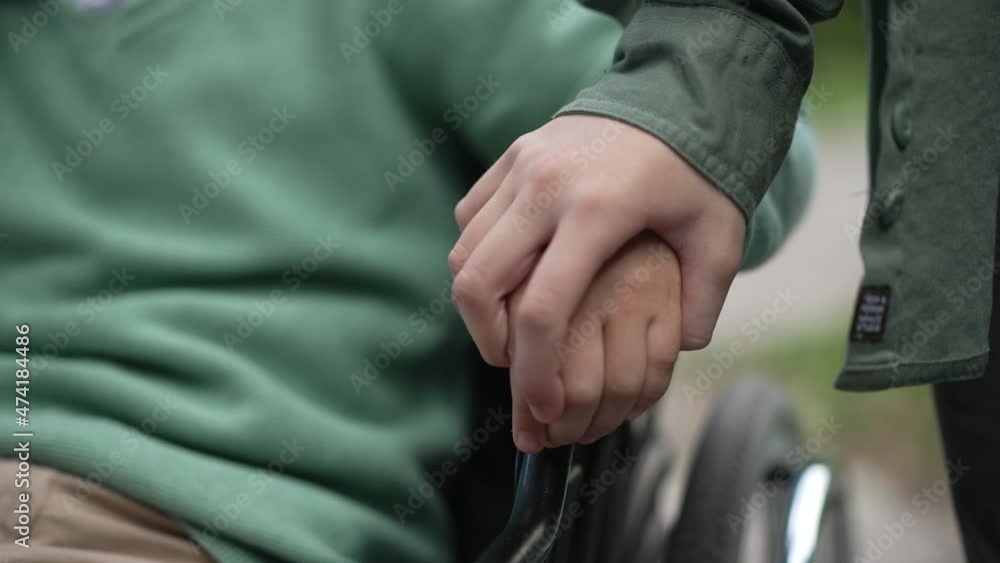 Close-up child hand on wheelchair armrest with unrecognizable girl touching palm of disabled friend in slow motion. Paralyzed Caucasian child outdoors supported by friend. Endorsement concept Close-up child hand on wheelchair armrest with unrecognizable girl touching palm of disabled friend in slow motion. Paralyzed Caucasian child outdoors supported by friend. Endorsement concept