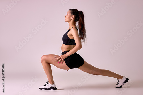 Fitness female doing lunges exercises for leg muscle workout training, active athlete lady is doing front forward one leg step lunge exercise, on pink studio background, side view