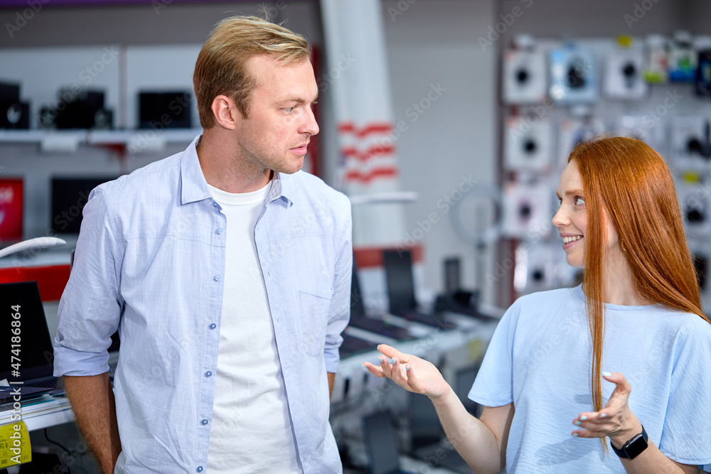 Obraz premium people in consumer electronics retail store talking, want to buy latest laptop, television and photo camera, look at each other, discussing. caucasina male and female in casual wear indoors in shop