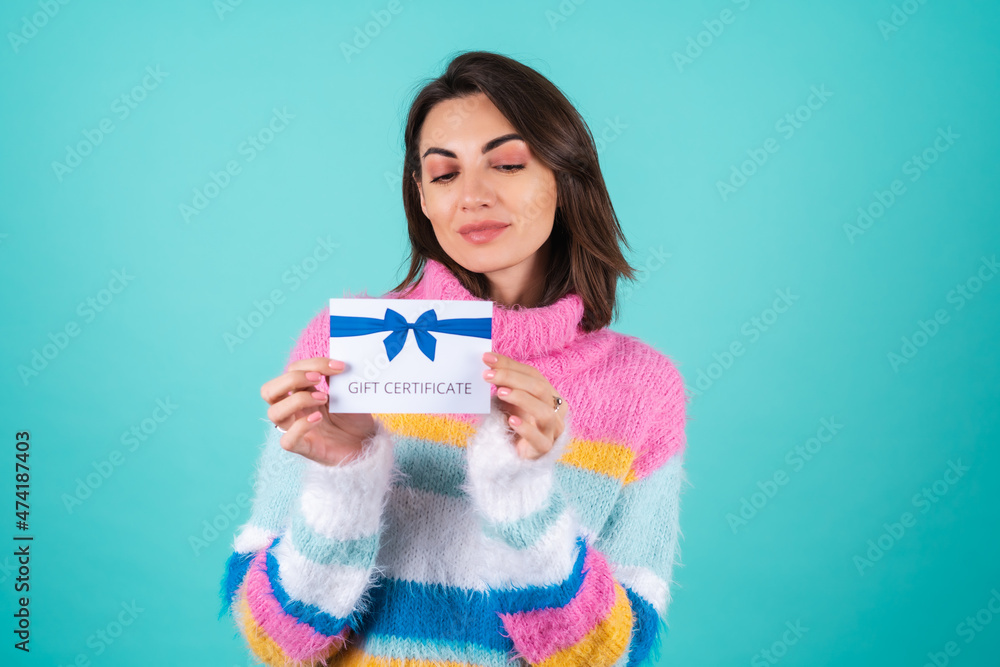 Young woman in a bright multicolored sweater on a blue background holding a gift certificate, cute smiling Young woman in a bright multicolored sweater on a blue background holding a gift certificate, cute smiling