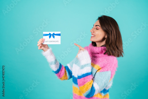 Young woman in a bright multicolored sweater on a blue background holding a gift certificate  smiling cute  pointing with a finger at a voucher