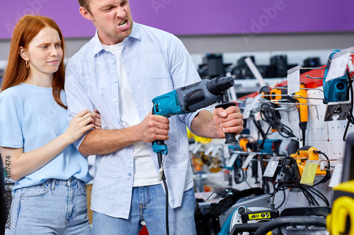 Irritated lady is upset by shopping with boyfriend choosing household tool instrument for repair. redhead woman and handsome guy in casual wear standing by shelves with tools, woman doesn't understand