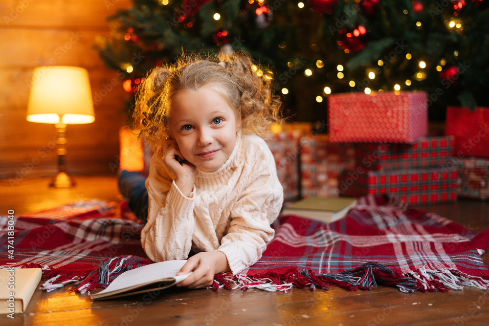 Portrait of happy cute little blonde curly child girl writing letter to Santa Claus lying on floor on background of Christmas tree and gift boxes, looking at camera, in room with festive interior. Portrait of happy cute little blonde curly child girl writing letter to Santa Claus lying on floor on background of Christmas tree and gift boxes, looking at camera, in room with festive interior.