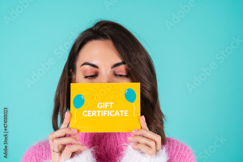 Young woman in a bright multicolored sweater on a blue background holds a gift certificate, smiles enthusiastically, beautiful makeup, covers the half of her face