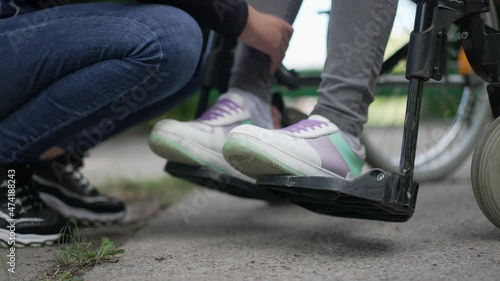 Legs of disabled teenager on wheelchair legrest with unrecognizable friend putting on sneakers in slow motion. Empathetic Caucasian teenage girl taking care of ill paraplegic friend outdoors in park photo