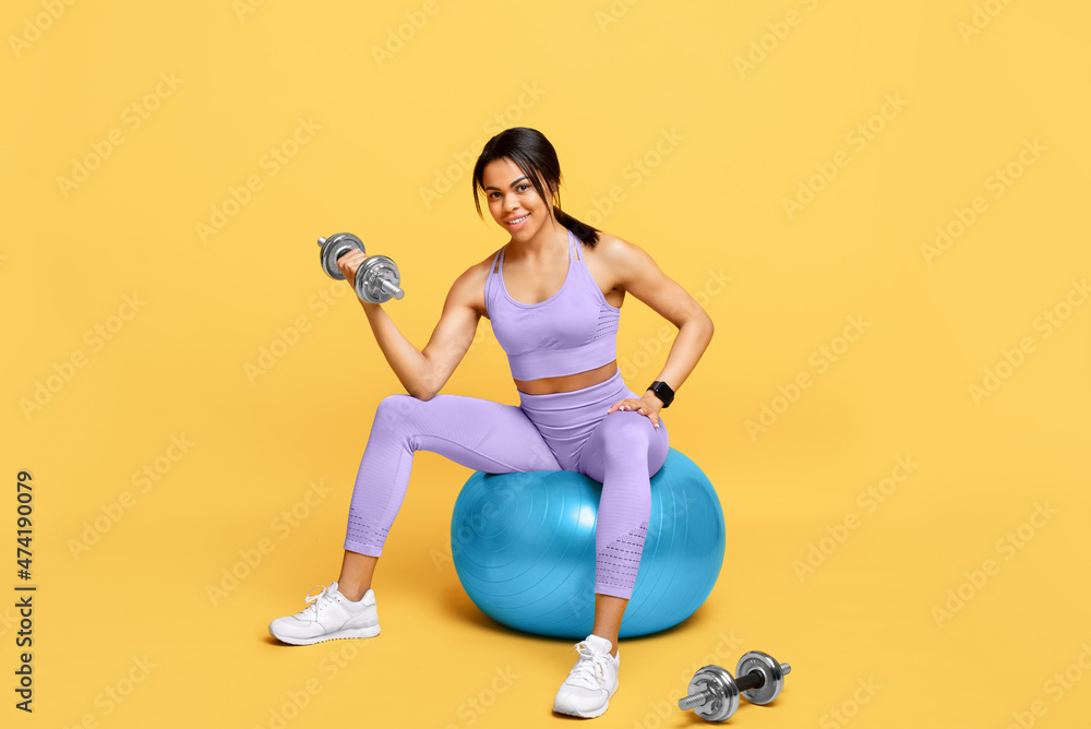 Fit african american woman sitting on fitness ball and exercising with barbells over yellow studio background Fit african american woman sitting on fitness ball and exercising with barbells over yellow studio background