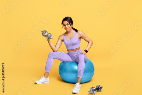 Fit african american woman sitting on fitness ball and exercising with barbells over yellow studio background